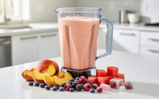 Close-up of a Vitamix blender pitcher with a creamy summer smoothie, surrounded by fresh peaches, berries, and watermelon on a sunlit kitchen counter.
